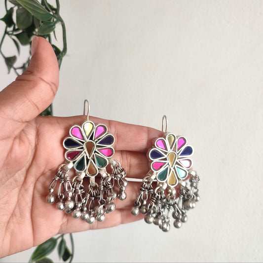 Colorful floral earrings with silver dangling parts held by a hand against a light background.