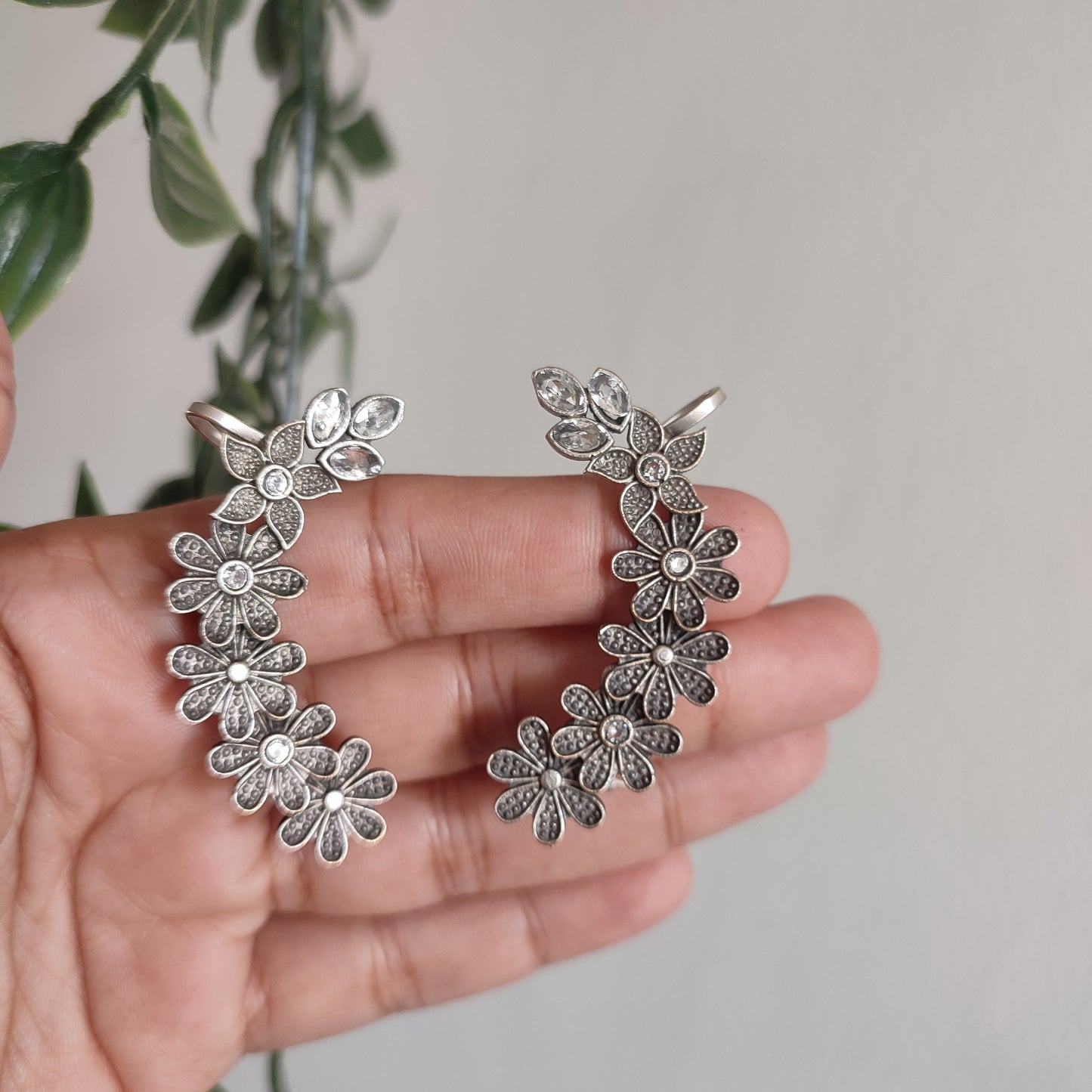 Silver floral earrings held in a hand with a blurred plant background